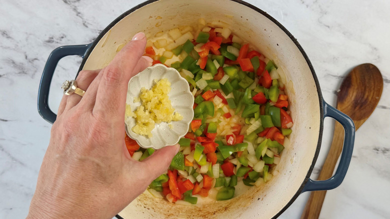 Hand adding small bowl of garlic to pot with onions and peppers