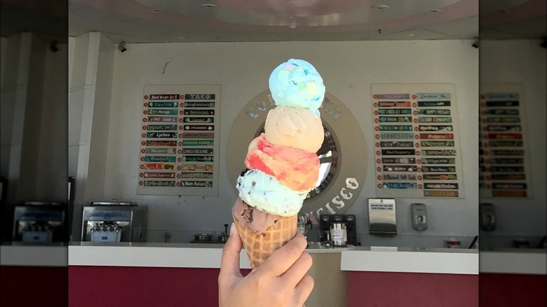 A hand holding a waffle cone layered with five scoops of ice cream in front of a wall full of flavor listings.