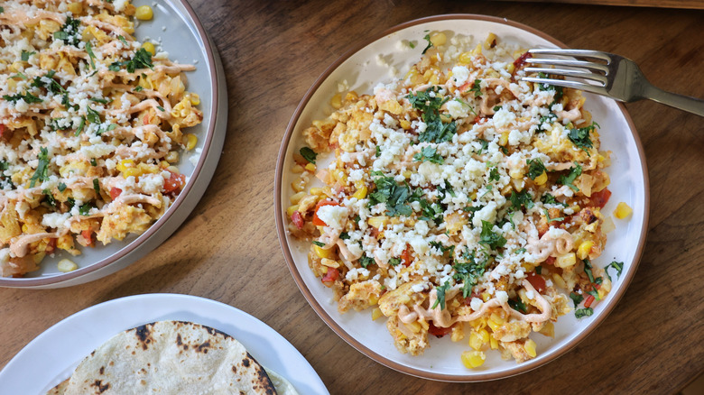 two plates of elotes-style breakfast scramble and a plate of charred corn tortillas on a wooden table