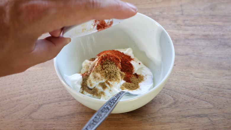 Hand pouring small dish of spices into bowl of sour cream with spoon in it