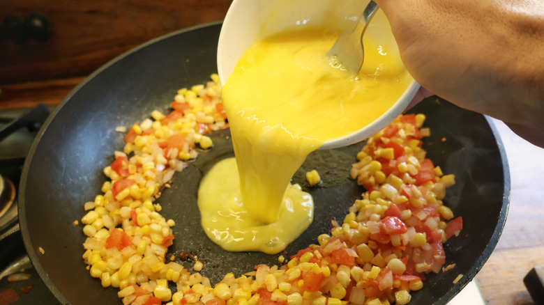 beaten eggs pouring from small bowl into center of skillet with corn, tomatoes, and onions