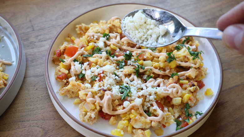 sprinkling a spoonful of crumbled cotija over a plate of elotes-style breakfast scramble