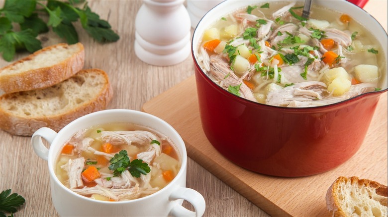 A pot of chicken soup with a cup of soup served next to slices of sordough and parsley 
