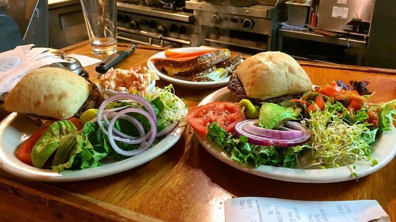 Two plates with burgers on ciabatta rolls and veggies on the side plus a plated sandwich in the back