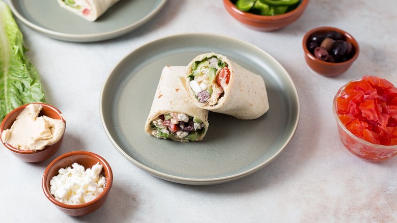 greek salad wrap on plate surrounded by various bowls of salad ingredients