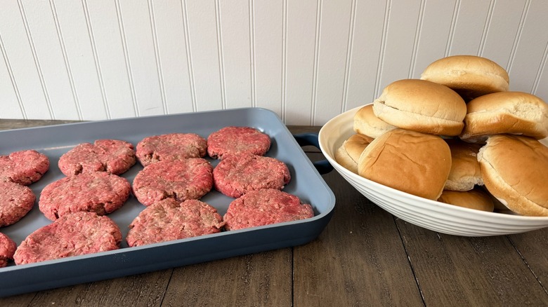 A pan with raw burger patties next to a bowl of burger rolls on a wooden table