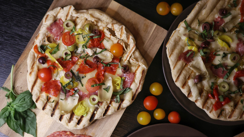 overhead shot of grilled pizzas topped with salami, olives, cheese, pickled pepper, artichokes, and chiffonade basil
