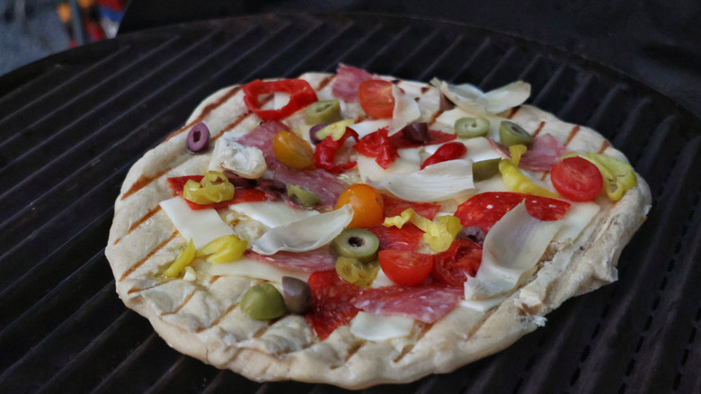 pizza on the grill topped with salami, olives, cheese, pickled peppers, and cherry tomatoes