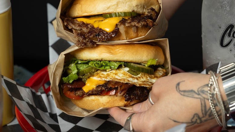 Hands holding cheeseburgers wrapped in brown paper