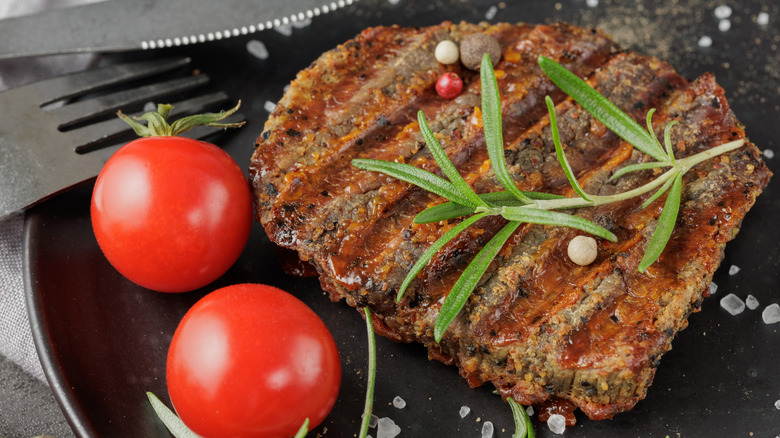 Cooked eye of round steak on black plate with two tomatoes and sprig of garnish.