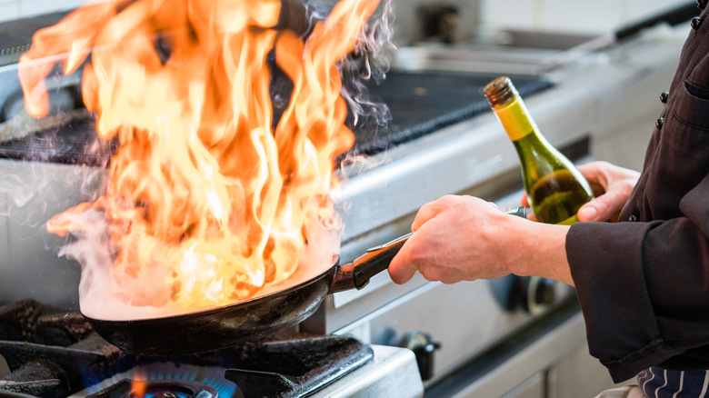 Chef holding a bottle while flambéing food in a pan