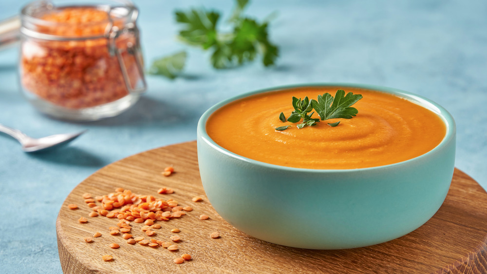 A bowl of red lentil soup. There are whole uncooked lentils scattered on the wooden cutting board next to it.