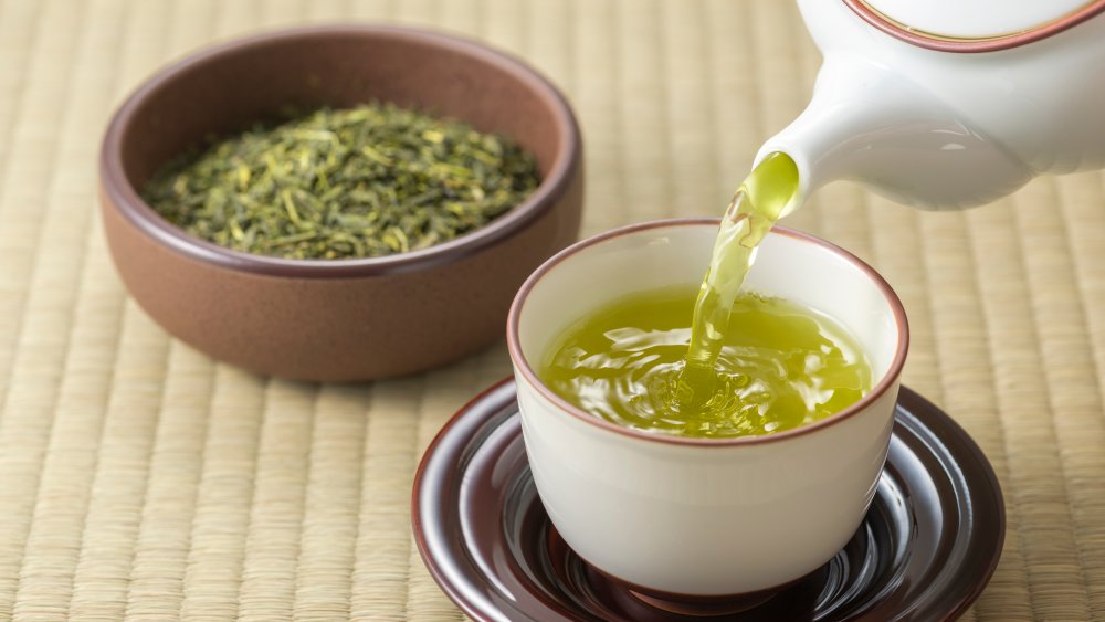 Green tea being poured into a cup with a bowl of green tea leaves in the background