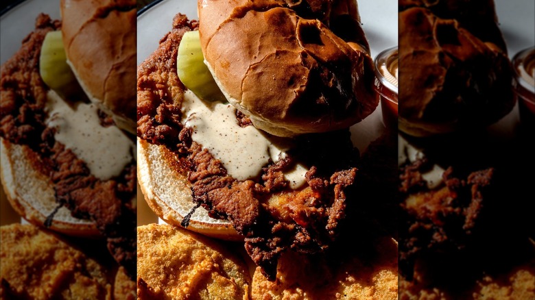 A close-up of a fried chicken sandwich with pickles and a creamy sauce on a toasted bun, served with crispy fried pickles on the side.