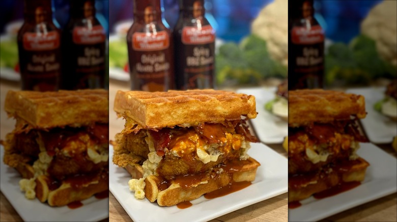 A chicken-and-waffle sandwich filled with fried chicken, macaroni and cheese, bacon, and a spicy BBQ sauce, served on a white plate.