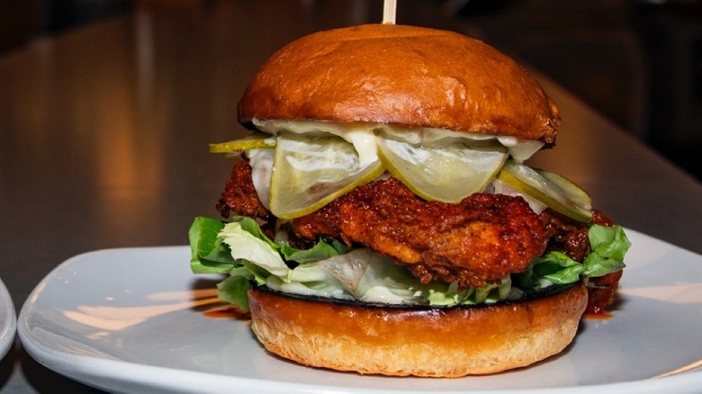 A fried chicken sandwich with pickles, lettuce, and sauce on a toasted bun on a white plate.