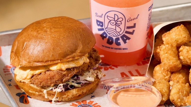 A fried chicken sandwich with slaw and pimento cheese sitting on a tray with tater tots, a dipping sauce, and a pink slushy drink.
