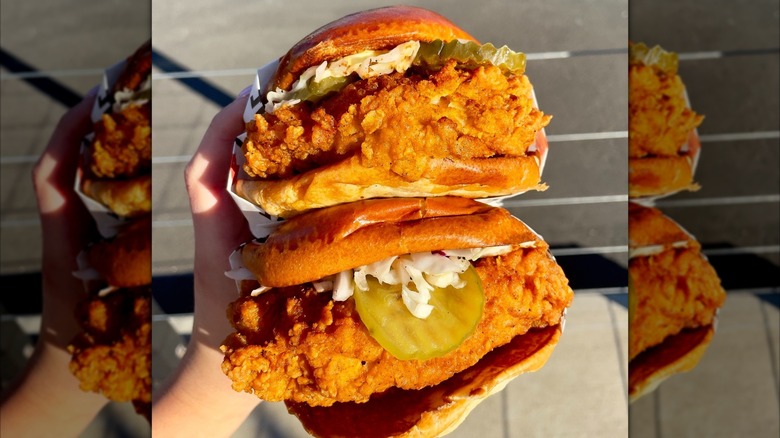 A hand holding two stacked fried chicken sandwiches with pickles, slaw, and toasted buns.