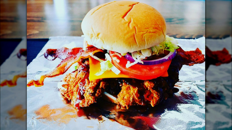 A fried chicken sandwich loaded with bacon, cheddar, lettuce, tomato, and red onion on a bun.