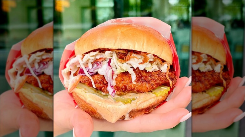 A fried chicken sandwich loaded with slaw, pickles, and a dripping sauce, wrapped in paper and held in a hand.