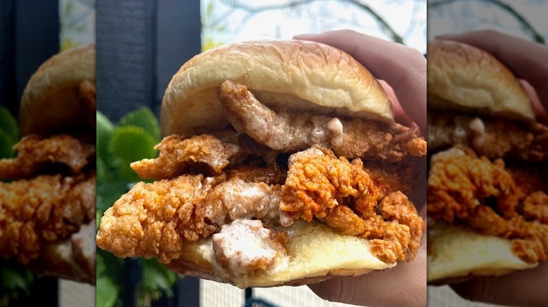 A hand holding a fried chicken sandwich filled with crispy chicken and creamy white sauce on a bun.