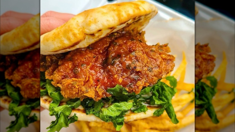 A close-up of a fried chicken sandwich with fried chicken and lettuce between two naan, and fries visible in the background.