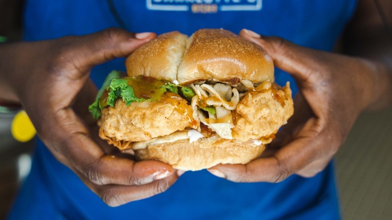 Hands holding a fried chicken sandwich with lettuce and sauce.