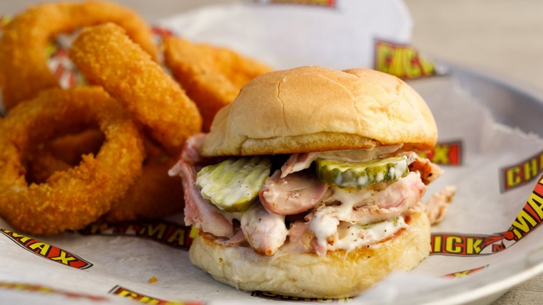 A close-up of a pulled chicken sandwich with pickles and a creamy sauce on a plate with onion rings