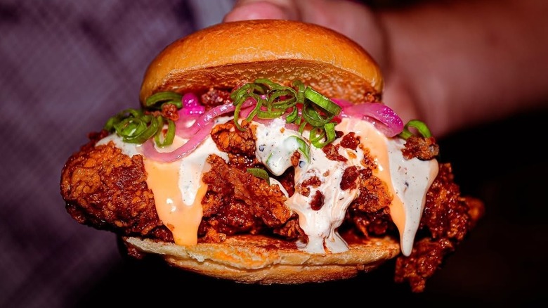A fried chicken sandwich topped with pickled onions, scallions, and drizzled with two different sauces.