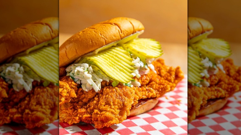 A close-up of a fried chicken sandwich with crinkle cut pickles and a slaw on red and white checkered service paper.