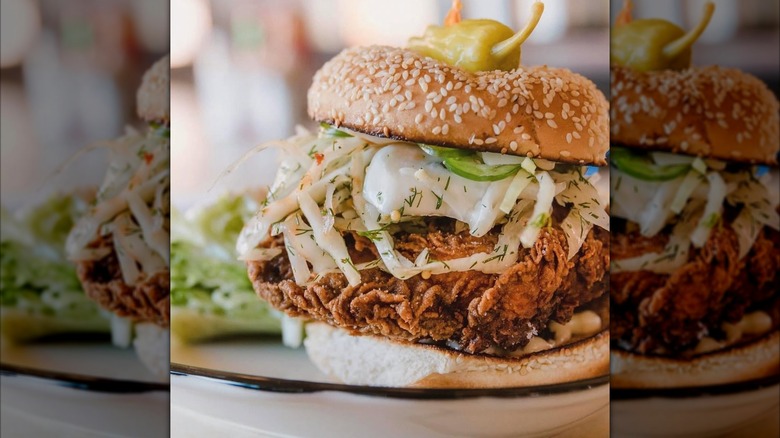 A close-up of a fried chicken sandwich on a sesame bun piled with dill slaw, pickles, and a pepperoncini on top.