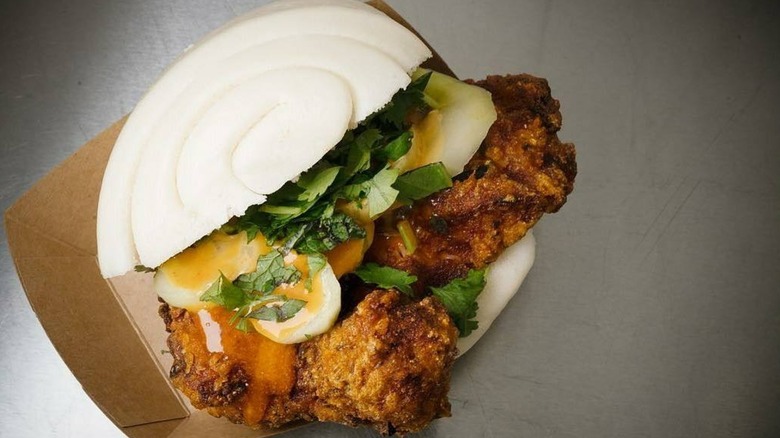 A bao bun filled with fried chicken, herbs, pickles, and sauce in a cardboard tray.