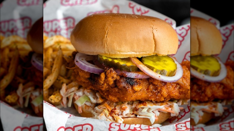 A fried chicken sandwich topped with pickles, red onion, and coleslaw, served on a bun with seasoned fries on the side