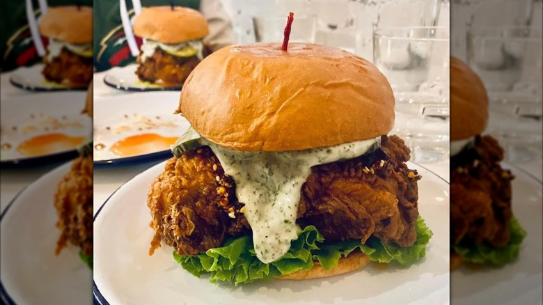 A fried chicken sandwich with lettuce, pickles, chili crunch, and a creamy aioli with herbs on a white plate