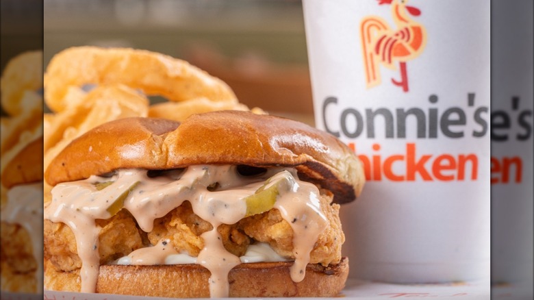 A fried chicken sandwich topped with a creamy sauce and pickles, served with onion rings and a drink that has the logo of Connie's Chicken