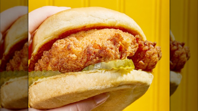 Someone holding a fried chicken sandwich with pickles on a soft bun in front of a yellow background