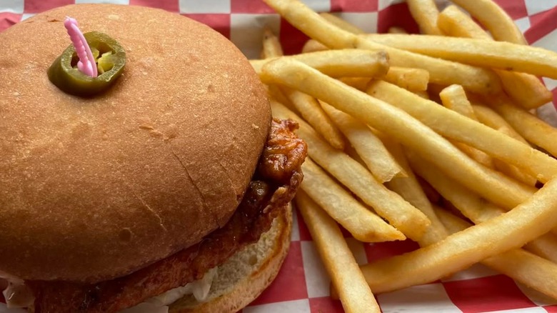 A fried chicken sandwich with coleslaw topped with a jalapeño slice and toothpick, served with a side of fries on a red-and-white checkered tray