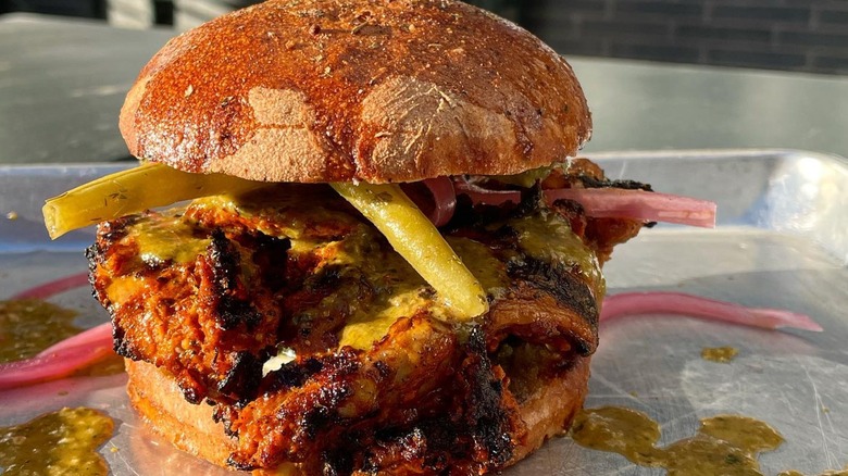 A charred fried chicken sandwich topped with green sauce, pickled onions, and peppers on a metal tray outside a restaurant
