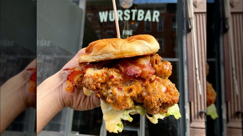 Someone holding a loaded fried chicken sandwich with bacon and sauce in front of a restaurant