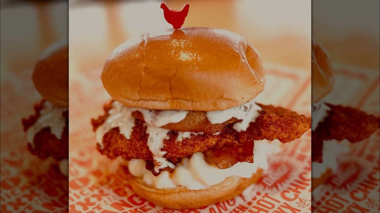 A fried chicken sandwich topped with white sauce, mac n' cheese, and an onion ring, secured with a red chicken-shaped pick