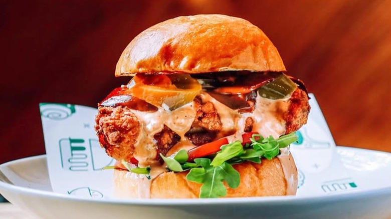 A fried chicken sandwich topped with sauce, pickled chilis, lettuce, and tomato on a plate