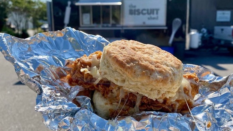 A chicken biscuit with cheese in a piece of aluminum foil in front of The Biscuit Shed food truck