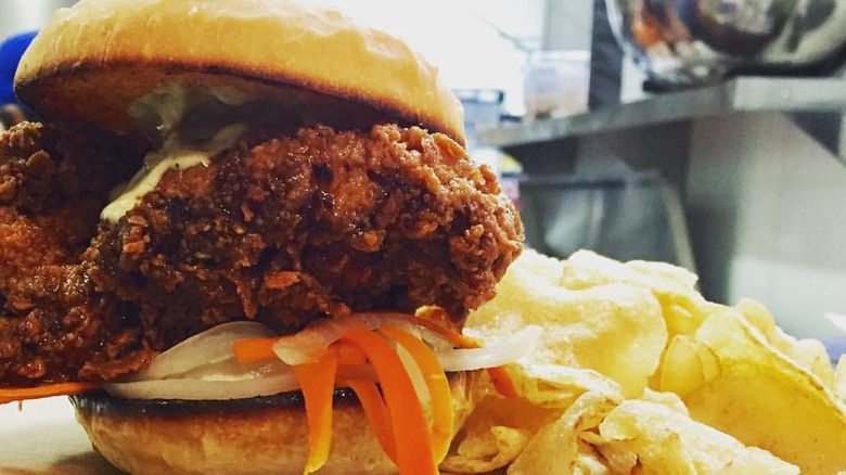 A fried chicken sandwich topped with pickled vegetables and served with potato chips