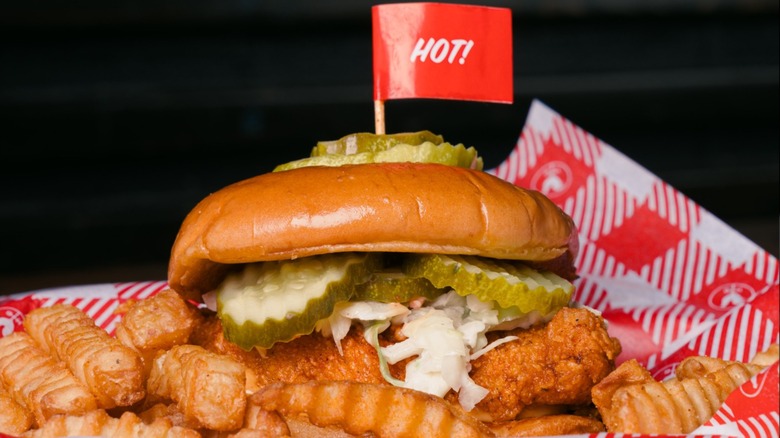 A fried chicken sandwich with coleslaw and pickles, topped with a small red flag that says HOT!, served with crinkle fries