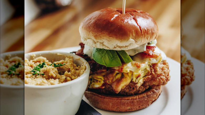A fried chicken sandwich topped with avocado, eggs, bacon, and sauce, served with a bowl of grits