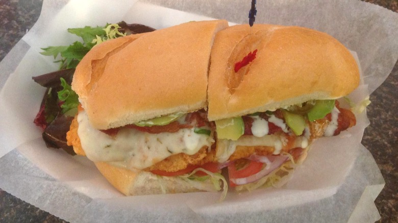 A fried chicken sub sandwich with lettuce, tomatoes, pickles, cheese, and sauce on a hoagie roll