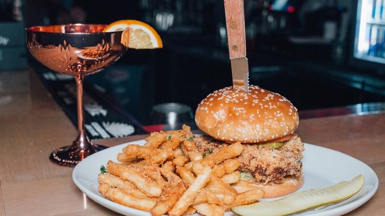 A fried chicken sandwich on a plate with seasoned fries, a pickle spear, and a knife stuck into the bun