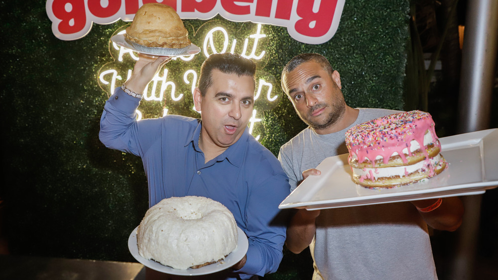 Buddy Valastro and Joe Ariel holding cakes in front of Goldbelly sign