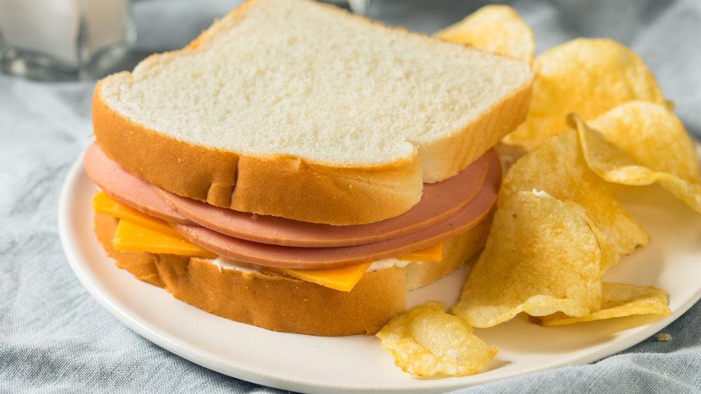 Bologna and cheese sandwich on white bread on white plate with potato chips on the side