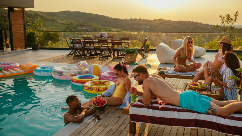 Handful of adults at sunset back-deck pool party with floats, beverages, and trays of fresh fruit